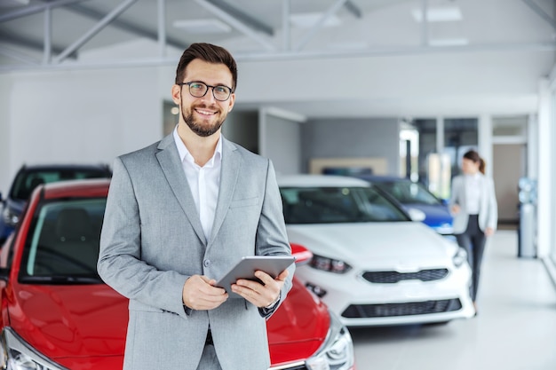 Smiling friendly car seller in suit standing in car salon and holding tablet. It's always pleasure to buy a car on a right place.