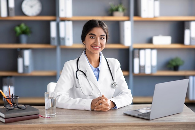 Smiling friendly young indian female doctor in white coat at workplace with laptop in clinic office interior