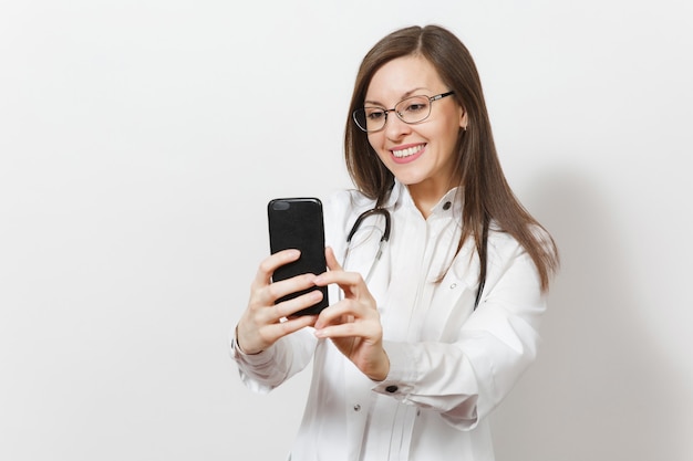 Smiling fun beautiful young doctor woman with stethoscope, glasses isolated on white background. Female doctor in medical gown doing selfie on mobile phone. Healthcare personnel, health, medicine conc