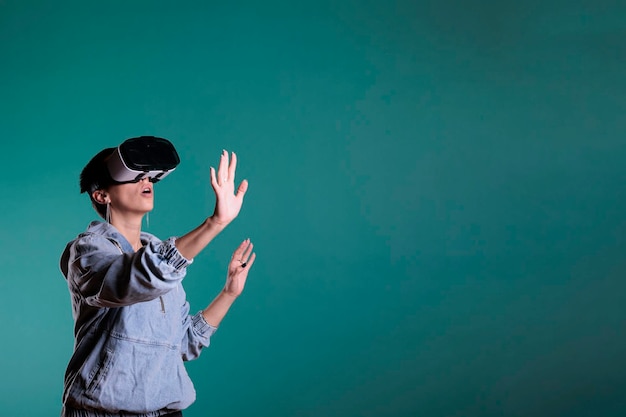 Smiling fun caucasian female wearing virtual reality headset with video interaction, standing over blue background. Confident woman playing videogames using vr technology with 3d tech simluation