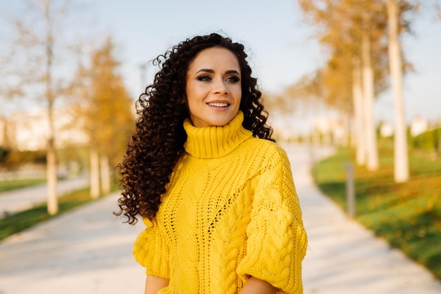 Smiling gentle smile girl in a bright sweater with curls of dark hair posing in the autumn park