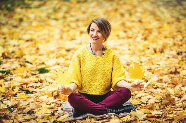 Smiling girl in autumn sitting on grass and holding two yellow leaves in hands