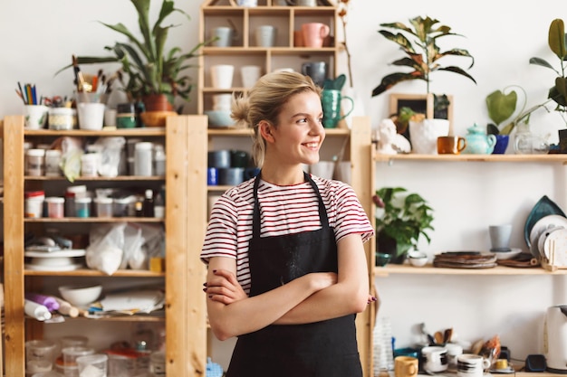 Smiling girl in black apron and T-shirt happily looking aside with handmade dishes on shelves on background at pottery studio