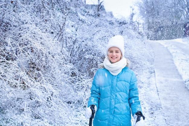 Smiling girl in blue winter clothes with Scandinavian walking sticks stands in the park.