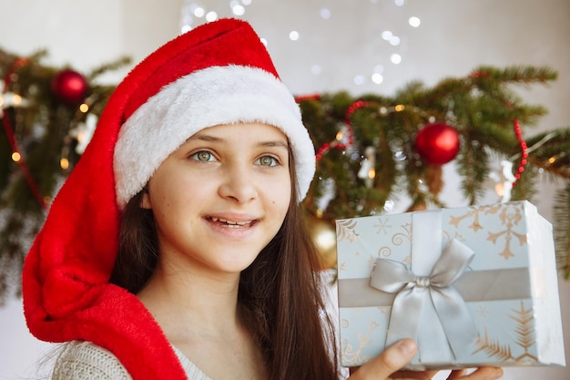 a smiling girl in a Christmas hat holds a gift box in her hands