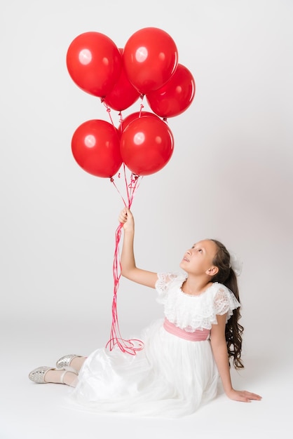 Smiling girl dressed in long white dress holding lot of party red balloons in hand looking up at balls