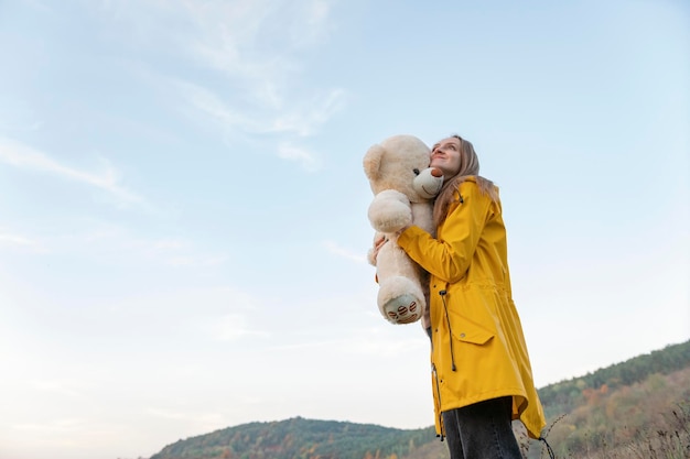 Smiling girl hugs teddy bear walks through mountainous terrain Young woman in yellow raincoat with soft toy outside