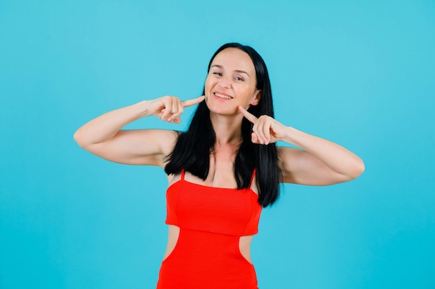 Smiling girl is holding forefingers on cheeks on blue background