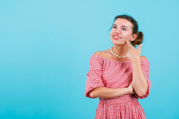 Smiling girl is looking up by holding forefinger on cheek on blue background