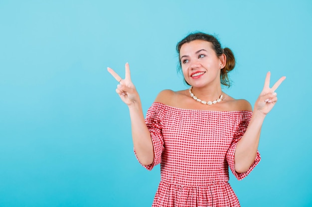 Smiling girl is looking up by showing victory gestures on blue background