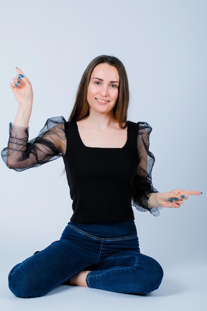 Smiling girl is pointing right and up with forefingers by sitting on floor on white background