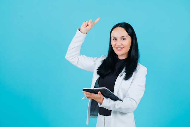 Smiling girl is pointing up with forefinger by holding tablet on blue background