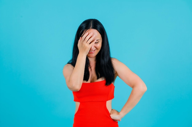 Smiling girl is putting hand on forehead and other on waist on blue background