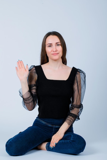 Smiling girl is showing hi gesture by sitting on floor on white background