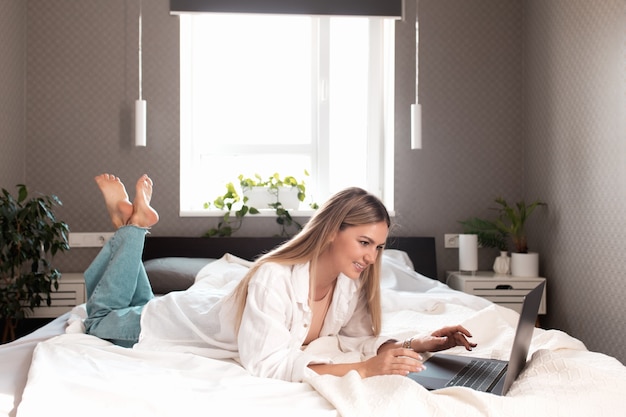 Smiling girl lays on the bed in bedroom and use laptop for remote work