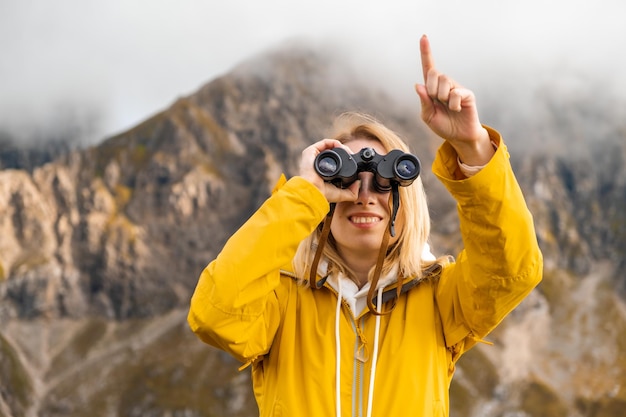 Smiling girl looking through binoculars and showing by finger in the distance with amazing mountains