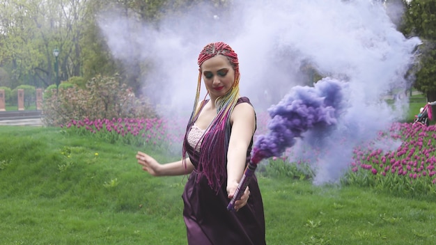 Smiling girl in purple satin dress with long multi-colored braids and eye-catching glitter makeup. Smoke of purple color covers the girl in the spring park