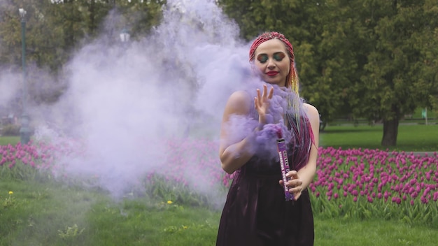 Smiling girl in purple satin dress with long multi-colored braids and eye-catching glitter makeup. Smoke of purple color covers the girl in the spring park