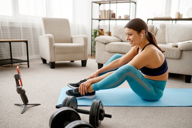 Smiling girl sits on the floor at home, online fitness training at the laptop. Female person in sportswear, internet sport workout, room interior