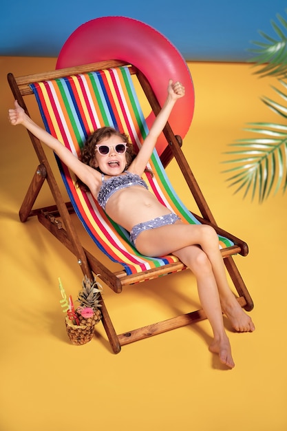 Smiling girl in swimsuit and sunglasses lying in rainbow deck chair with hands raised up emotionally and sunbathing