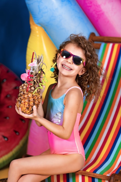 Smiling girl in swimsuit and sunglasses sitting in rainbow deck chair holding pineapple cocktail with colorful straws and showing thumb up and sunbathing