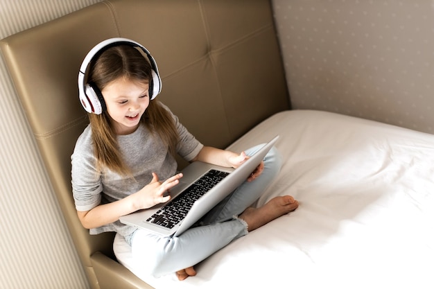 Smiling girl in wireless headphones using her laptop on the bed at home