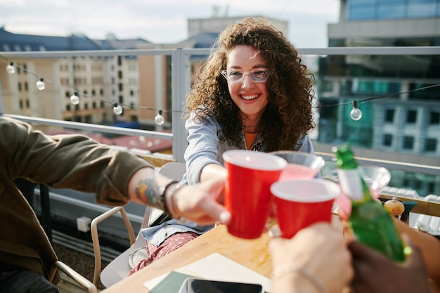 Smiling girl with dark long curly hair toasting with drinks of her friends