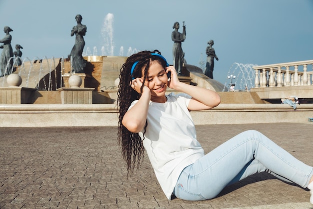 A smiling girl with dreadlocks in headphones is sitting on the floor in the park A happy young woman relaxing with headphones enjoying music Space for copyinggeneration z