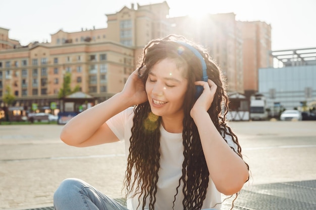 A smiling girl with dreadlocks in headphones is sitting on the floor in the park A happy young woman relaxing with headphones enjoying music Space for copyinggeneration z