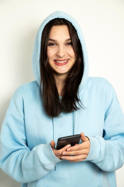 Smiling girl with phone Cute model in a blue sweater with a hood on a white background reads sms