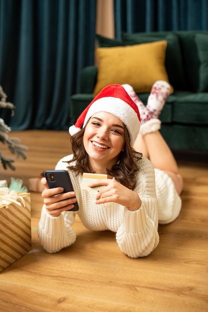 Smiling girl with smartphone and credit card near Christmas tree. Online shopping on new year. She buying presents