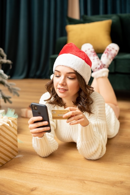 Smiling girl with smartphone and credit card near Christmas tree. Online shopping on new year. She buying presents