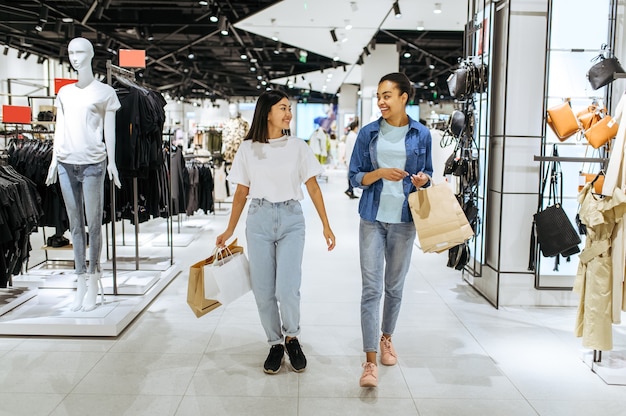 Smiling girlfriends with cardboard bags walks in clothing store. Women shopping in fashion boutique, shopaholics, shoppers with purchase