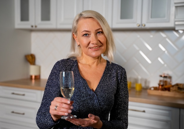 smiling grandmother in beautiful festive dress standing in stylish kitchen holding champagne glass 