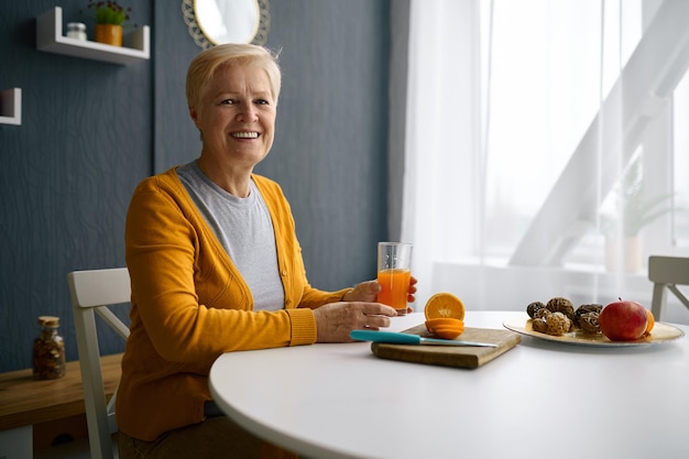 Smiling grey-haired woman holding glass cup with orange juice. Freshly picked oranges on table