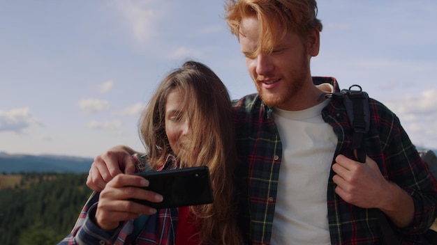 Smiling guy and girl looking video on smartphone screen during hike in mountains Happy couple of friends using mobile phone at vacation Handsome man hugging woman with cellphone outdoor