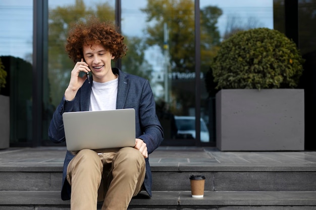 Smiling guy programmer working outdoors using laptop talking on smartphone drinking coffee