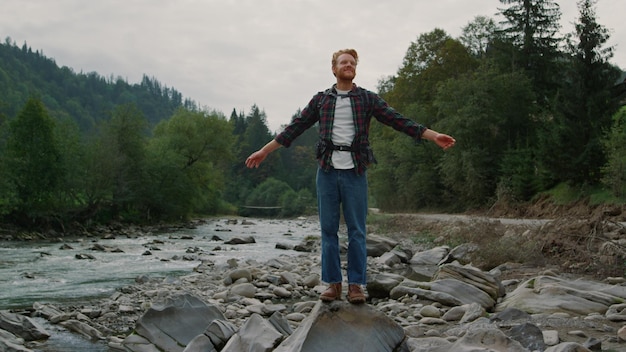 Smiling guy standing on rock at river shore Male tourist with backpack raising hands in air Positive hiker feeling freedom at mountain river landscape Redhead man screaming outdoor