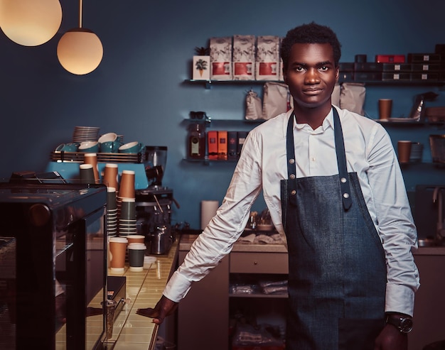 Smiling handsome African American barista in uniform holds cup of coffee while standing in hiscoffee shop.