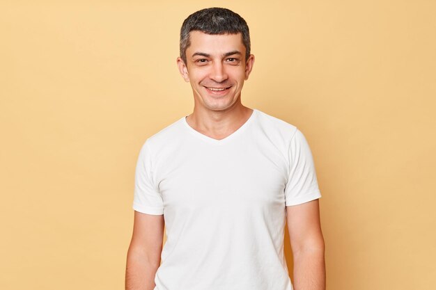 Smiling handsome man wearing white casual tshirt standing isolated over beige background looking at camera being in good mood expressing positive emotions