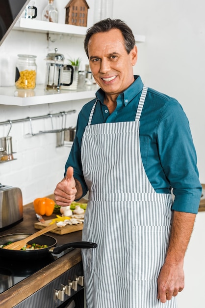 Smiling handsome mature man cooking vegetables on frying pan in kitchen and showing thumb up