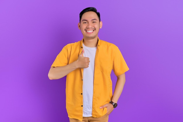 Smiling handsome young Asian man showing thumb up gesture looking at camera isolated on purple background