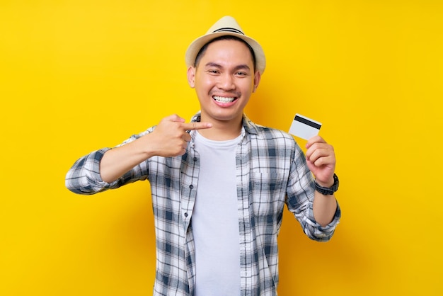 Smiling happy Asian ethnicity young man 20s wearing casual clothes hat pointing finger at credit bank card isolated on yellow background studio portrait People lifestyle concept