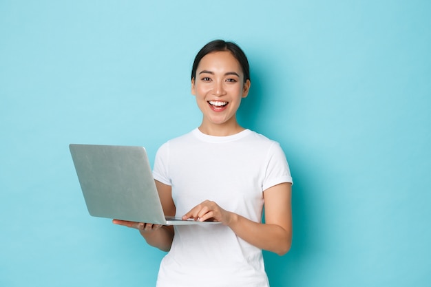 Smiling happy, beautiful asian woman using laptop while standing over light blue wall, express cheerful attitude, working on project or shopping online