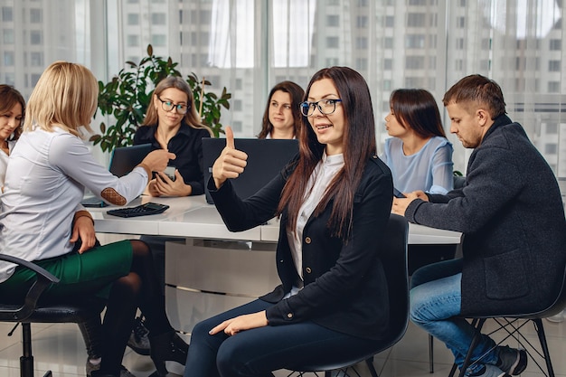 Smiling happy brunette woman wearing glasses sitting on a chair in the office lifting a finger up surrounded by her colleagues immersed in work. Concept of job