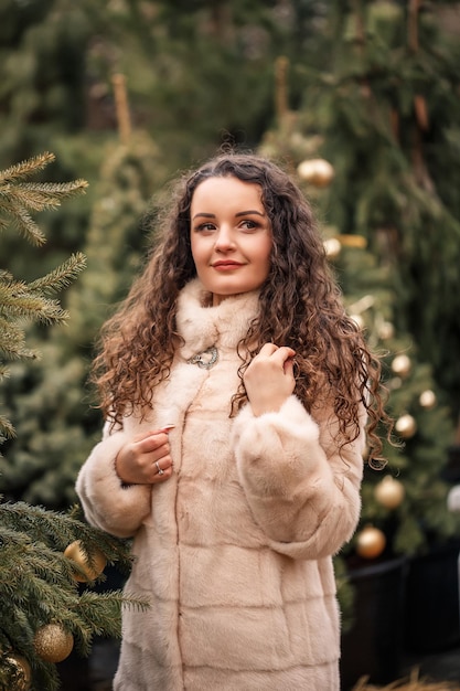 Smiling happy curlyhaired woman in a fur coat strolls through the Christmas tree fair Waiting for Christmas and New Year