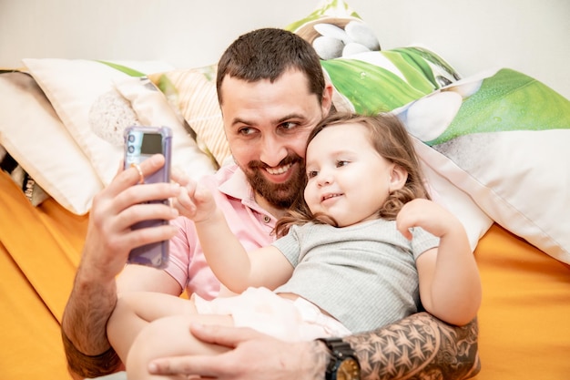 Smiling happy father with his little cute daughter looking at smartphone screen taking making selfie portrait photography sitting on couch having fun together happy family concept