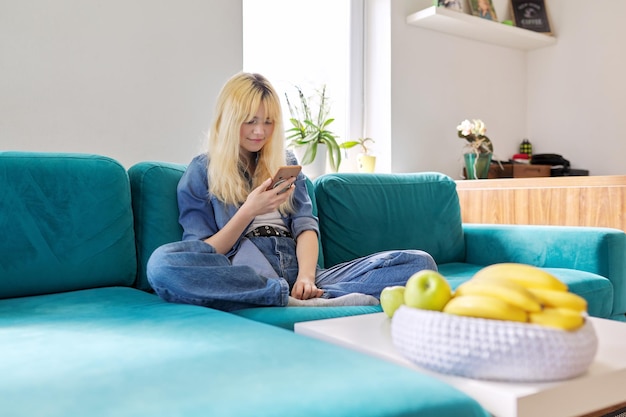 Smiling happy female teenager sitting at home on couch looking at smartphone screen