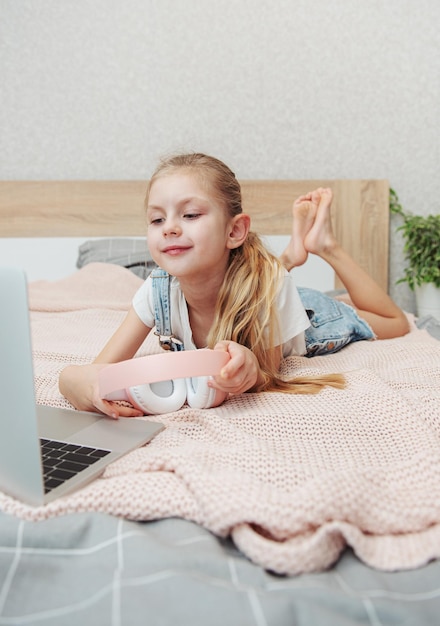 Smiling happy little kid girl using laptop in wireless headphones in bed at home.