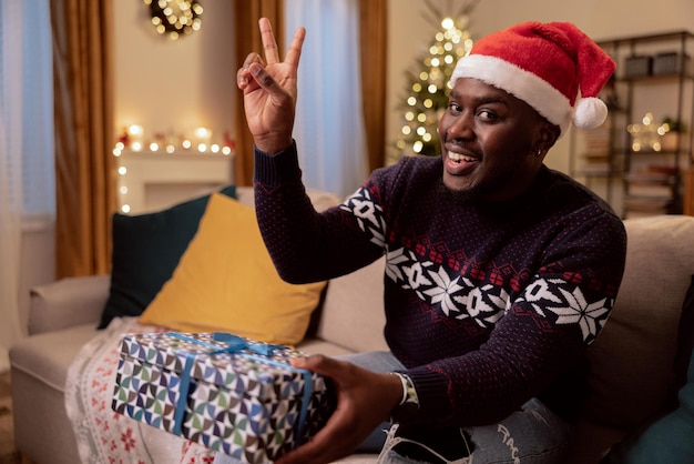 A smiling happy man is sitting on the couch during christmas and holding a wrapped gift box